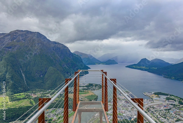 Fototapeta  Rampestreken lookout point on the trail to Nesakla summit in Norway.  Cloudy sky, grey water of Romsdalsfjord, dark mountains, town of Andalsnes down there