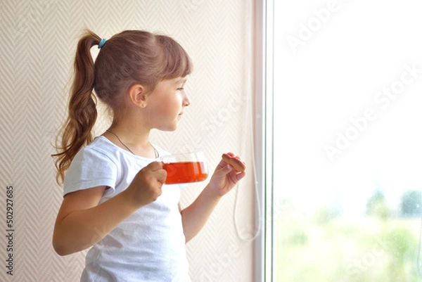 Fototapeta A girl in a white T-shirt holds a cup of tea and a liver in the form of a red heart standing by the window, watching. delicious sweet snack. safe at home, self-isolation during a pandemic.