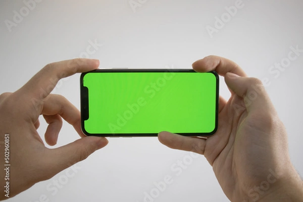 Obraz Holding an iPhone in hand. Green Screen on the phone. Mock-up of an iPhone. Apple product in hand. Vertical tilt of the phone.