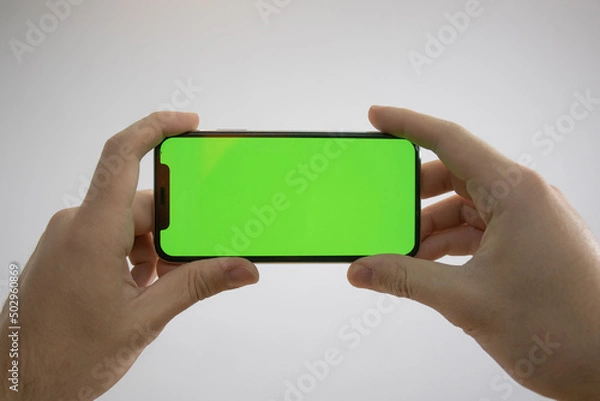Obraz Holding an iPhone in hand. Green Screen on the phone. Mock-up of an iPhone. Apple product in hand. Vertical tilt of the phone.