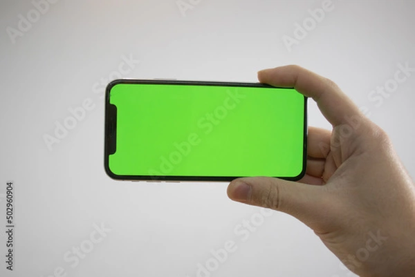 Obraz Holding an iPhone in hand. Green Screen on the phone. Mock-up of an iPhone. Apple product in hand. Vertical tilt of the phone.