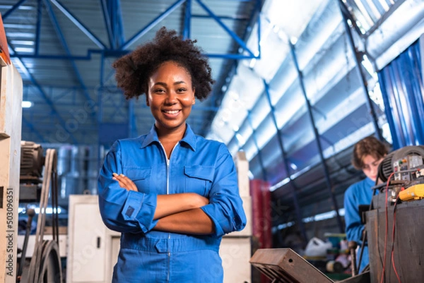 Fototapeta Engineer african woman wearing safety helmet working and checking machine  automotive part warehouse factory.Happy female  worker looking at the camera.
