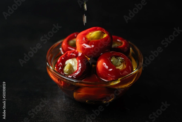 Fototapeta Red chili peppers stuffed with ricotta and capers in oil, smeared with drops of pouring oil at long exposure, on a dark background