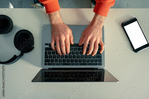Obraz Laptop, young man checking his social media accounts, the phone next to him has a white screen, headphones and listening to music, workstation