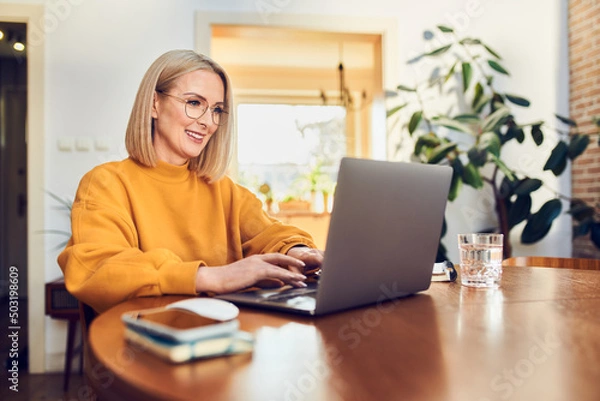Fototapeta Portrait of middle aged woman sitting at dinning with laptop working at home