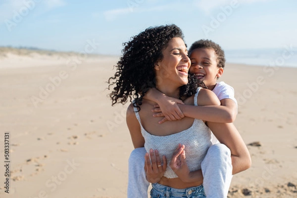 Fototapeta Joyful mother and son spending time on beach. African American family walking, laughing, playing, riding on back. Leisure, family time, parenthood concept