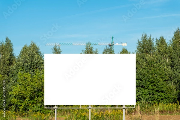 Obraz Blank white billboard for advertisement in front of trees. There is also tower crane in the background. Idea of city development.