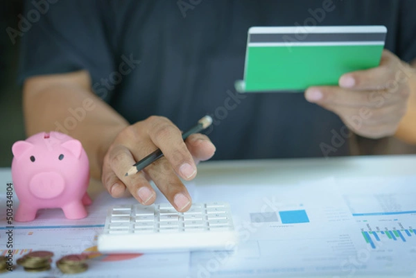 Obraz Man hand pressing calculators and holding saving account passbook, piggy bank with coin.Save money and buy house concept.
