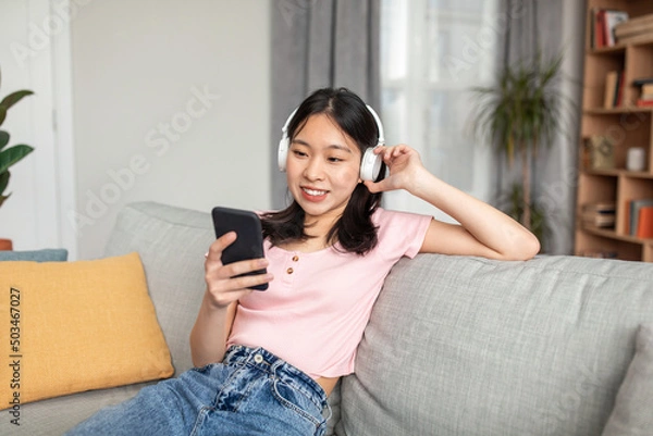 Obraz Cheerful korean lady in headset relaxing on couch, listening to music or watching movie on smartphone at home