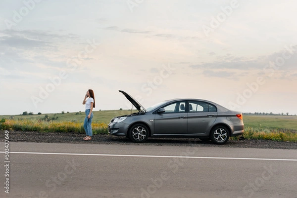 Fototapeta A young girl stands near a broken car in the middle of the highway during sunset and tries to call for help on the phone and start the car. Waiting for help. Car service. Car breakdown on the road.
