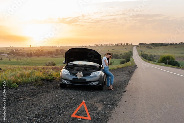 Fototapeta A frustrated young girl stands near a broken-down car in the middle of the highway during sunset. Breakdown and repair of the car. Waiting for help. Car service. Car breakdown on road.