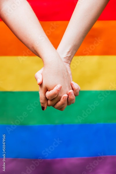 Fototapeta Close up of two woman's hands holding each other with the LGBTQ rainbow flag in the background. Symbol for homosexual community for pride month and the celebration day of sexual and love diversity