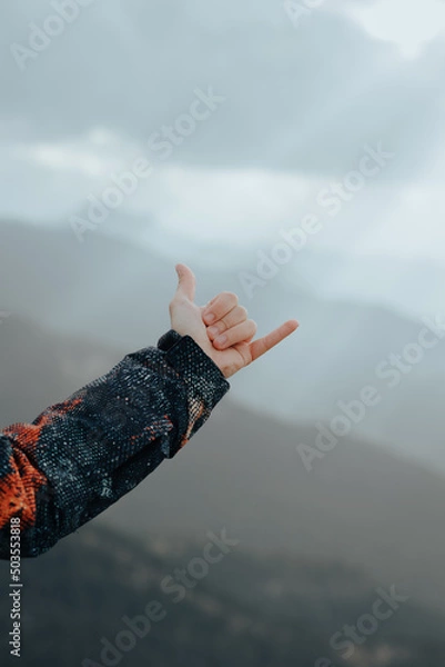 Fototapeta Unrecognizable male hand wearing a coat making the hang loose sign on top of a mountain with peaks in the background