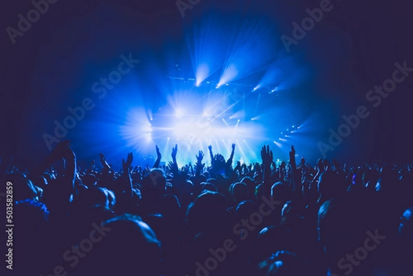 Fototapeta A crowded concert hall with scene stage lights in blue tones, rock show performance, with people silhouette, on dance floor air during a concert festival