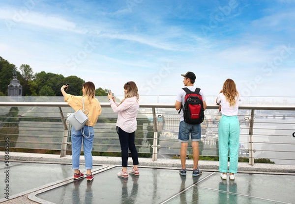 Fototapeta Young tourists on the glass bridge in Kyiv. View of the Dnieper river with rocks, green hills. Urban city life.