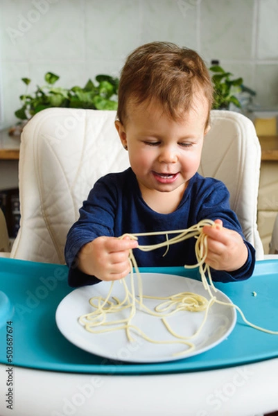 Obraz Little cute baby learning to eat himself at the kids table in the kitchen. It feels happy.