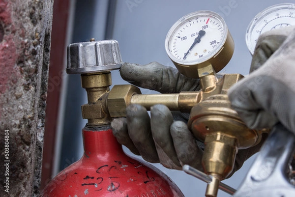 Obraz Red bottle for storing liquefied gas. Gas cylinder. Close-up plan of Men's hands in gloves screwing the reducer on the gas cylinder. Concept for content about fuel, gas or welding.