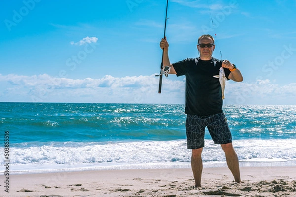 Fototapeta An elderly white caucasian man, 60 years old, holds a fishing rod and a freshly caught Whiting fish (pollock, Theragra chalcogramma) against the backdrop of the Atlantic Ocean in Florida. Copy space