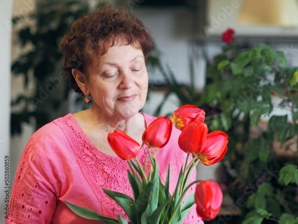 Fototapeta Charming old woman grandmother in bright clothes with a bouquet of tulips at home.