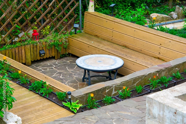 Fototapeta Patio design. Patio made of natural stone and composite boards. Round table with a hidden hearth. Benches around the table. Flower beds around