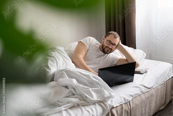 Obraz smiling man using Tablet Computer is lying on a bed in the morning. work from home. freelancer.