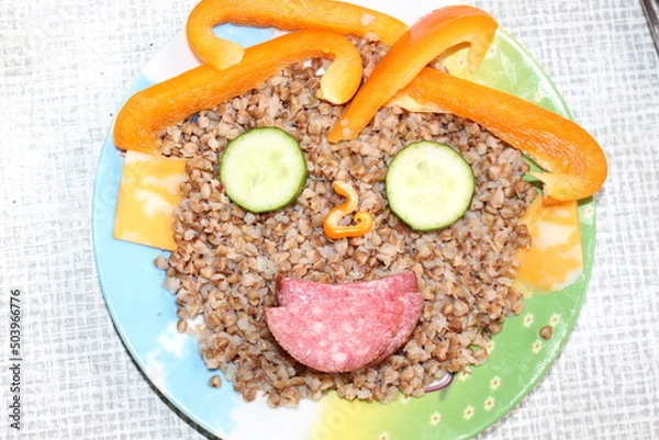 Fototapeta Breakfast for a child is decorated in the form of a funny face in a plate. Buckwheat, pepper, cucumber, cheese, sausage are good for the body