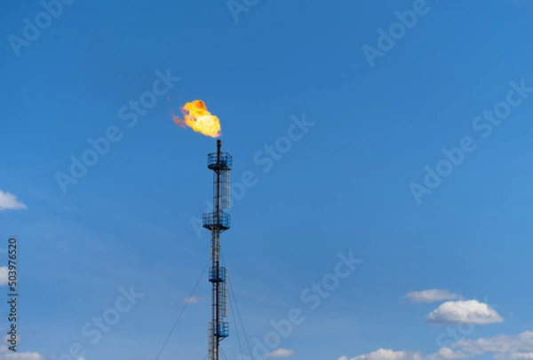 Fototapeta A gas flare at a field in the Perm region (Russia) against a blue sky. Gas tower with a bright pillar of fire. Flaring of associated petroleum gas. Photo minimalism.