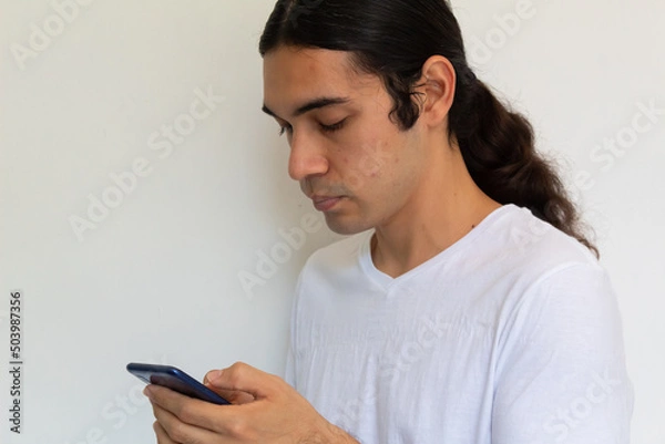 Obraz portrait of a non binary hispanic man with long hair on a white background using a cell phone