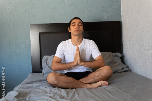 Obraz Hispanic non binary long-haired man doing yoga and meditating on his bed