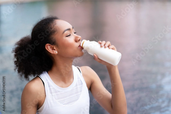 Fototapeta Young black woman with curly hair drinking fresh milk while sitting beside the swimming pool after workout for burning. Need protein, calcium, and good fat to build her muscles and control food, sugar