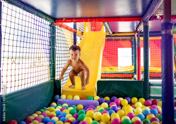 Fototapeta The kid plays in the playroom and rolls down the slide into the pool with small balls