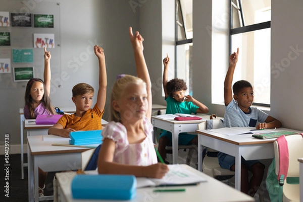 Fototapeta Multiracial elementary school students raising hands while sitting at desk in classroom