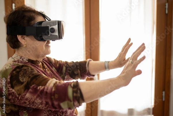 Fototapeta Caucasian aged mature woman exploring or playing in virtual augmented reality with vr goggles headset in the Metaverse. Futuristic world concept