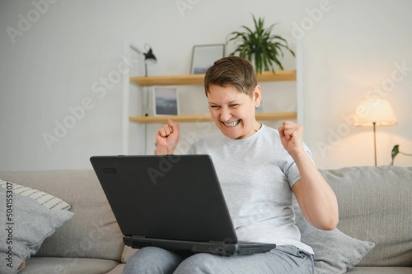 Fototapeta Excited mature woman looking at laptop screen, reading good news in message, celebrating online lottery win, rejoicing success, overjoyed older female sitting on couch at home, using computer.