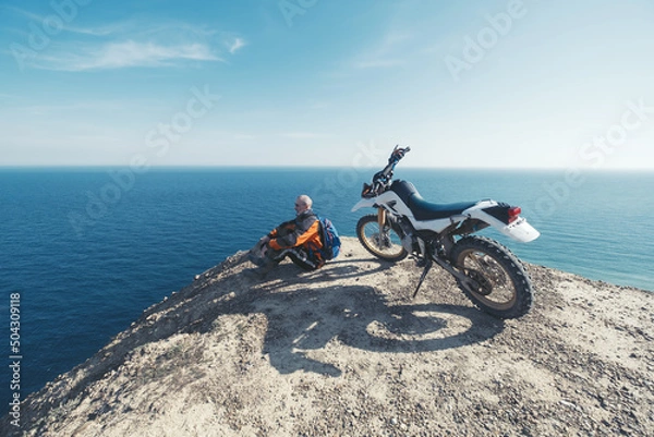 Fototapeta Elderly man resting in enduro motorcycle trip on mountain cliff with amazing sea skyline view