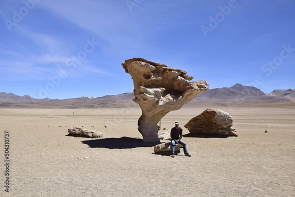 Fototapeta A man is sitting next to the famous stone tree rock formation (Arbol de Piedra) in the Siloli desert. Bolivia