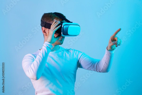 Fototapeta Modern young man experiencing virtual reality glasses in futuristic neon illuminated studio