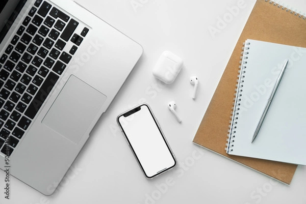 Fototapeta Mobile phone with blank white screen on desktop. Business concept template: laptop, mobile phone, wireless headphones, notepad and pen.