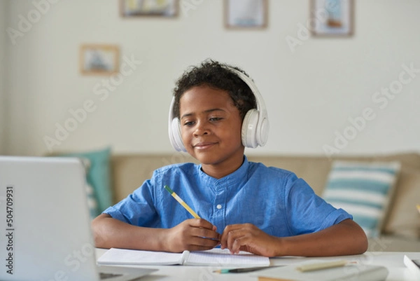 Fototapeta Content curious African-American boy in wireless headphones using laptop while talking to tutor via video conferencing app