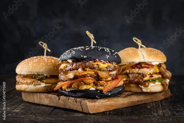 Obraz A set of craft juicy burgers on wooden cutting board on a smoky black background. trung on wooden skewers. Tasty grilled cheeseburgers with beef cutlet, tomato, cheese and lettuce. 