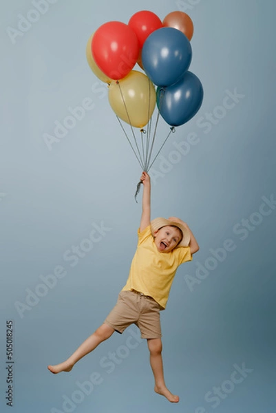 Fototapeta A funny kid in shorts and a straw hat is flying with colorful balloons on an isolated white background