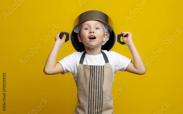 Fototapeta Funny little chef in an apron holding a pot on his head on an isolated yellow background. the concept of helping mom in the kitchen