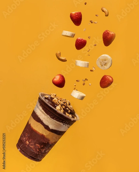 Fototapeta Acai cup, layers of granola, ground peanuts, and pieces of fruits and nuts flying in the air, yellow background.