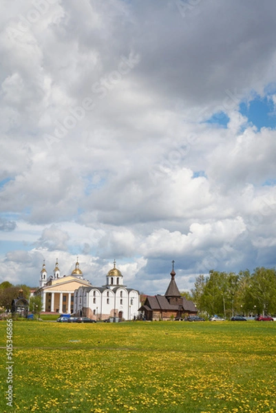 Fototapeta Spring urban landscape of a small town. In the foreground is a field of yellow dandelions, in the background are several churches. Most of the frame is occupied by an expressive cloudy sky.