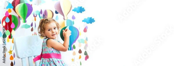 Fototapeta Caucasian curly haired girl in a beautiful blue dress on a white isolated background sits on a highchair. Multi-colored balloons, raindrop made of paper. Education, creativity, child care. Copy space