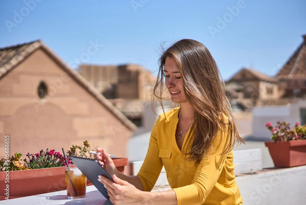 Fototapeta young happy woman working at her computer on the terrace of a Spanish café wearing a yellow blouse on a hot summer day
