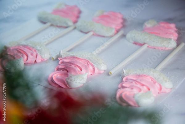 Fototapeta Meringue in the form of pink Christmas hats with a pompom and a white lapel on a stick lie on white parchment in two rows. High quality photo