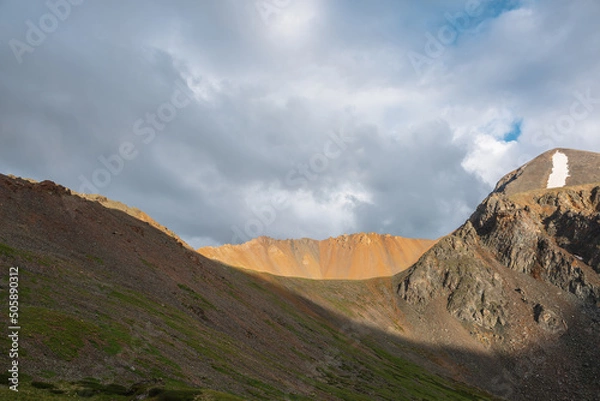 Fototapeta Dramatic alpine landscape with sunlit wide sharp mountain ridge under overcast sky at changeable weather. Atmospheric mountain scenery with large sharp rocks on ridge top in sunlight under cloudy sky.