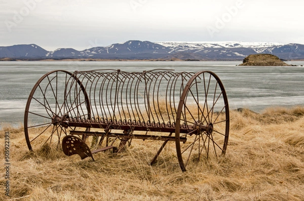 Fototapeta Rusty piece of agricultural equipment on a grassy slope on the shore of frozen lake Myvatn in Iceland