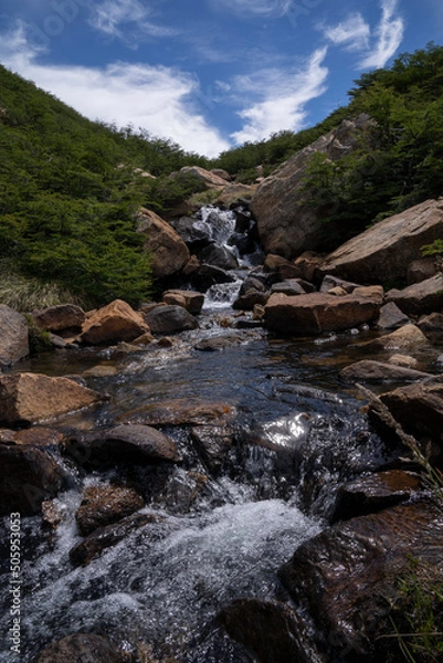 Fototapeta View of the waterfall flowing across the rocky mountain and forest in a summer sunny day. 